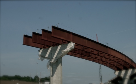 cropped photograph of a highway bridge under construction, showing heavy I-beams looming beyond the column support