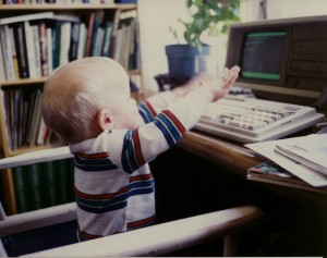 a blonde 12-month old boy seated on a chair, reaching up and pecking at the keyboard to an early 1990's personal computer